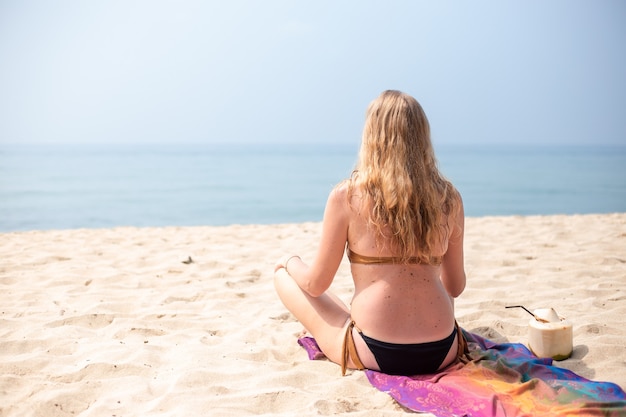 Una donna con lunghi capelli biondi siede nella posizione del loto in riva al mare sabbioso in Thailandia. Viaggi e turismo.