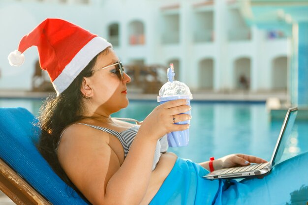 Una donna con un cappello da Babbo Natale beve un cocktail su una sedia a sdraio con un laptop vicino all'hotel