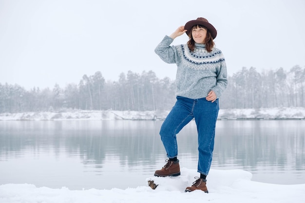 Una donna con un cappello e un maglione marroni si trova sulle rive di un lago e di una foresta innevati