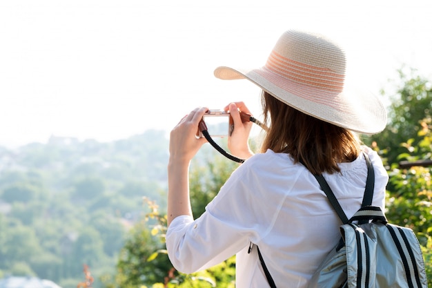 Una donna con un cappello e una felpa bianca con uno zaino viaggia e scatta una foto