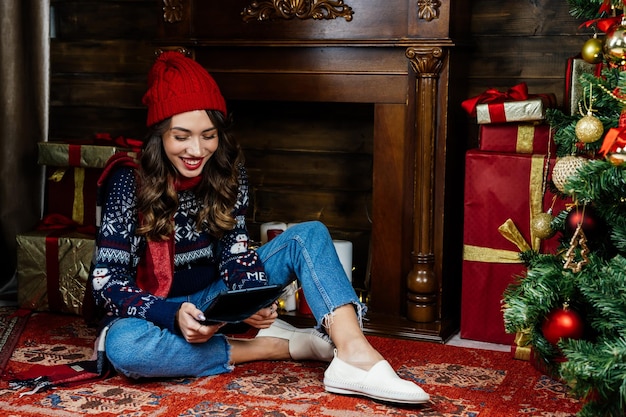 Una donna con un cappello e una sciarpa rossi ride delle informazioni contenute nel tablet Una bella bruna siede sotto un albero di Natale sullo sfondo delle decorazioni di Capodanno