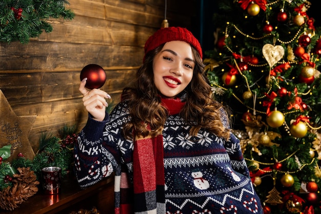 Una donna con un cappello rosso e una sciarpa ride Una bella bruna sta accanto al caminetto sullo sfondo di un albero di Natale e di decorazioni natalizie