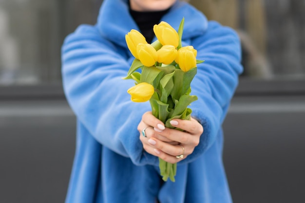 Una donna con un cappotto blu tiene in mano un mazzo di tulipani gialli