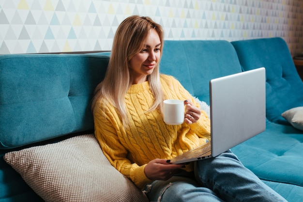 Una donna con un laptop in un maglione giallo è seduta sul divano