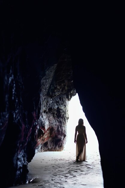 Una donna con un lungo abito di chiffon esce da una grotta sulla spiaggia. Ha le spalle in controluce