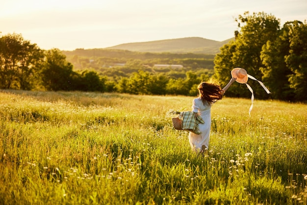 una donna con un vestito leggero e un cappello in mano corre lontano nel campo durante il tramonto