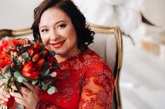 Una donna con un vestito rosso si siede su una sedia e tiene un mazzo di rose rosse e fragole all'interno.