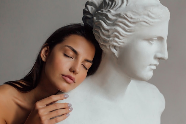 Una donna con un viso e un corpo ben curati si trova vicino al busto di una scultura di una donna