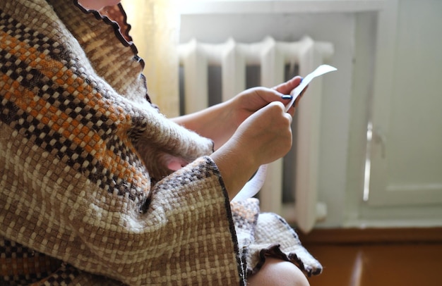 Una donna con una coperta calda si siede a casa davanti alla batteria e tiene in mano una bolletta del gas Concetto di crisi energetica Foto orizzontale