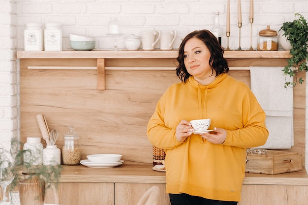 Una donna con una felpa arancione beve caffè in cucina a casa.