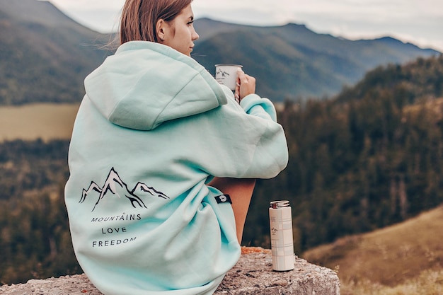 Una donna con una felpa con cappuccio blu con una stampa si siede su una pietra e tiene in mano una tazza di tè. Accanto c'è un thermos. Sfondo di pittoresche montagne rocciose.