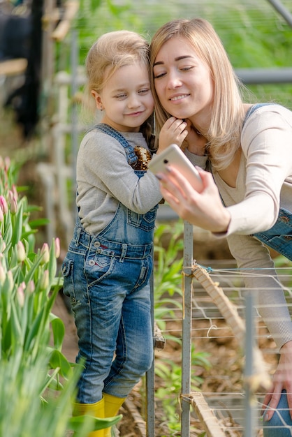 Una donna con una figlia piccola si fa un selfie in una serra fiorita in primavera.