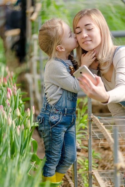 Una donna con una figlia piccola si fa un selfie in una serra fiorita in primavera.