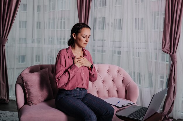 Una donna con una maglietta rosa si siede sul divano e conduce una consultazione a distanza usando un laptop