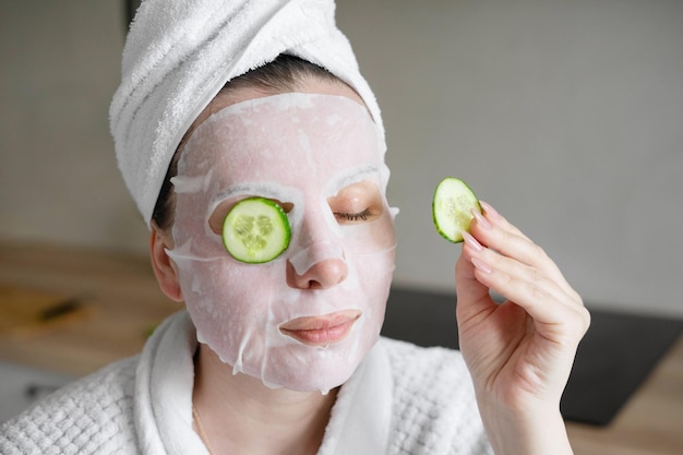 Una donna con una maschera cosmetica in tessuto e con un asciugamano in testa si mette delle fette di cetriolo sugli occhi La cura mattutina in cucina