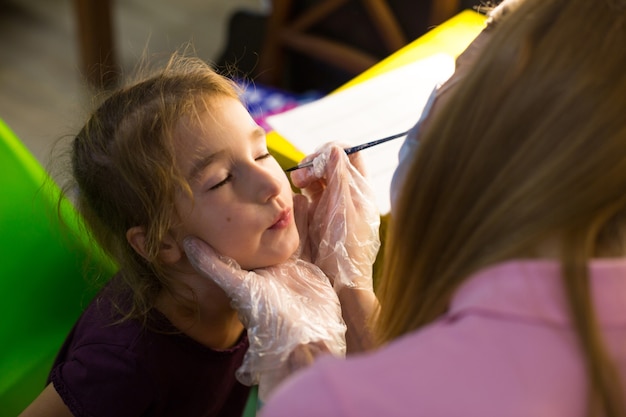 Una donna con una maschera medica disegna un motivo aquagrim sul viso di un bambino nello studio davanti a uno specchio con lampade. Divertimento per i bambini: colorazione del viso. Russia, Mosca, 15 agosto 2020