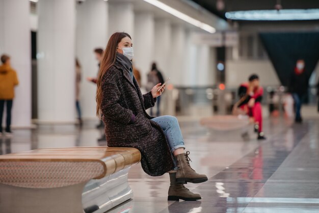 Una donna con una maschera medica è seduta al centro della piattaforma della metropolitana con uno smartphone e aspetta un treno. Una ragazza con una mascherina chirurgica sta mantenendo le distanze sociali in metropolitana.