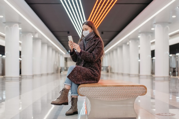 Una donna con una maschera medica è seduta al centro della piattaforma della metropolitana con uno smartphone e sta facendo un selfie. Una ragazza con i capelli lunghi e una mascherina chirurgica sta mantenendo le distanze sociali in metropolitana.