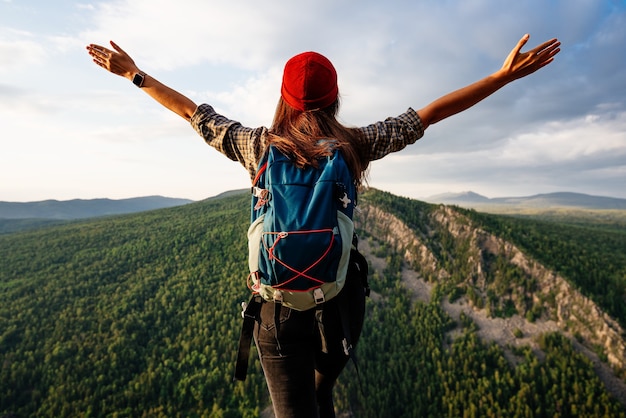 Una donna con uno zaino si erge sulla cima di una montagna con le mani alzate e ammira la bellezza di una valle di montagna, una vista posteriore. La ragazza viaggia in posti bellissimi. Tramonto in montagna