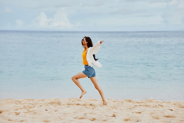 Una donna corre lungo la spiaggia in abiti estivi sulla sabbia in una maglietta gialla e pantaloncini di jeans camicia bianca capelli volanti vista sull'oceano