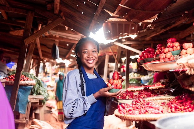 Una donna d'affari africana che indossa un grembiule colorato che vende pomodori e verdure in un tipico mercato africano locale