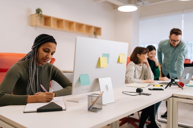 Una donna d'affari afroamericana concentrata che scrive appunti e usa il computer seduta al lavoro in un grande