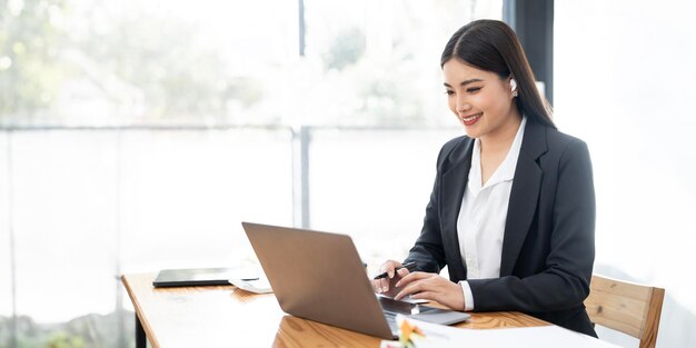 Una donna d'affari allegra che lavora al portatile in ufficio, un'asiatica felice, una bellissima donna di affari in abito formale che lavora sul posto di lavoro.