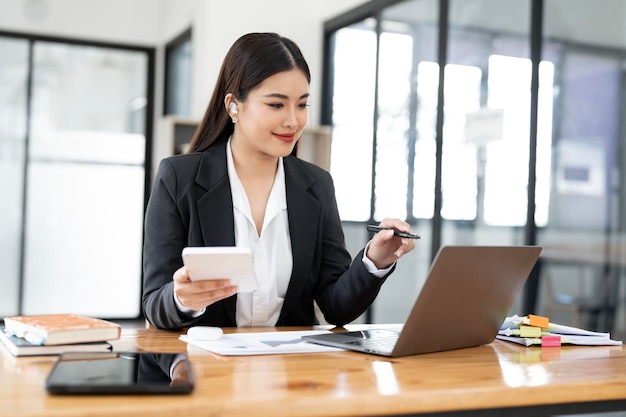 Una donna d'affari allegra che lavora con un portatile e una calcolatrice in ufficio, una bella donna d'affaires asiatica felice in abito formale che lavora sul posto di lavoro.