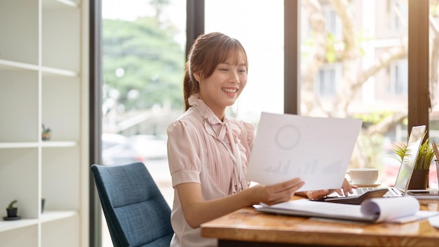 Una donna d'affari asiatica felice sta esaminando un rapporto finanziario che analizza i dati di vendita alla sua scrivania