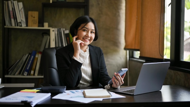 Una donna d'affari asiatica sorridente siede alla sua scrivania nel suo ufficio con un telefono in mano