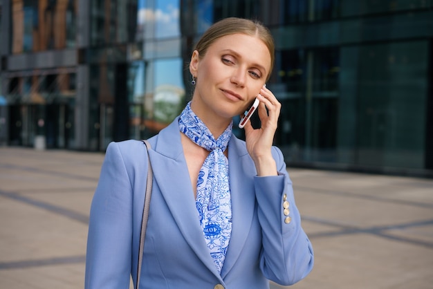 Una donna d'affari bionda vestita con una giacca rigorosa sta parlando al telefono sullo sfondo di un grattacielo.