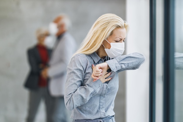 Una donna d'affari con maschera protettiva tossisce al gomito in ufficio durante la pandemia del virus corona.