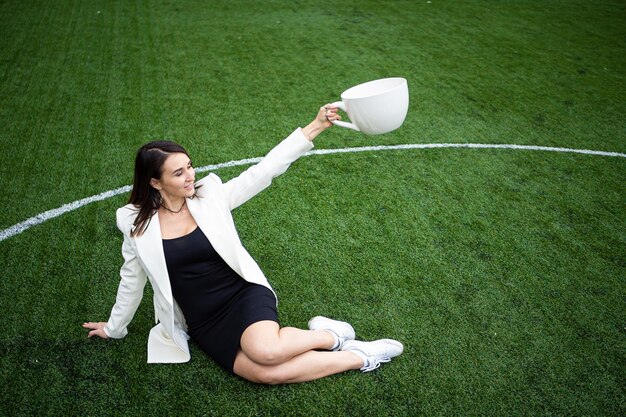 Una donna d'affari con una grande tazza seduta su un prato verde nel parco Il concetto di un ufficio
