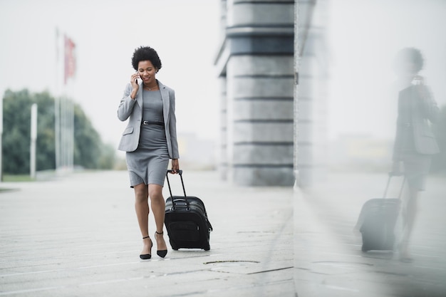 Una donna d'affari nera di successo che cammina con i suoi bagagli e usa lo smartphone fuori dall'aeroporto.