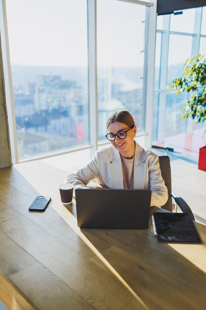 Una donna d'affari positiva con una giacca è seduta a un tavolo e lavora su un laptop in un ufficio luminoso Una giovane donna europea è seduta a un tavolo Freelance e lavoro a distanza Stile di vita femminile moderno