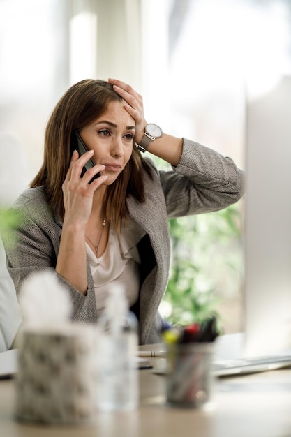 Una donna d'affari preoccupata che sembra stressata mentre parla sullo smartphone e lavora al computer nel suo ufficio.