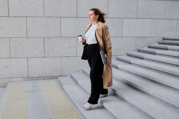 Una donna d'affari professionista cammina con fiducia verso l'edificio degli uffici bilanciando il suo portatile e sorseggiando il suo caffè mattutino