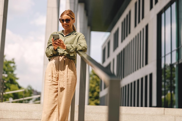Una donna d'affari sorridente con gli occhiali da sole sta usando il telefono mentre si trova sullo sfondo di un edificio moderno