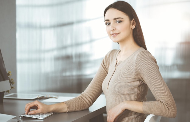 Una donna d'affari vestita casual e amichevole sta lavorando sul suo computer, mentre era seduta alla scrivania in un ufficio soleggiato. Concetto di successo aziendale