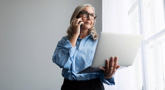 Una donna dai capelli grigi d'affari maturi di aspetto europeo parla al telefono con un computer portatile in lei