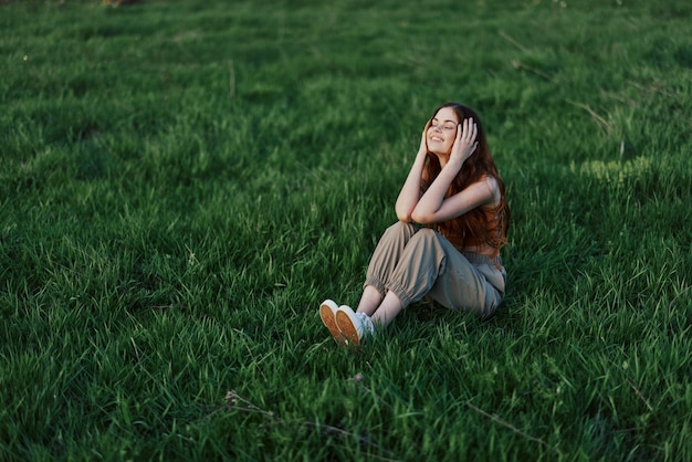 Una donna dai capelli rossi con lunghi capelli spazzati dal vento siede all'aperto sull'erba del parco e sorride alla luce del tramonto che le illumina il viso Il concetto di armonia con la natura all'aperto