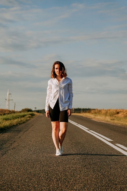 Una donna dai capelli rossi con una camicia bianca cammina lungo la strada tra i campi in una giornata di sole estivo. Una gita fuori porta. Foto verticale