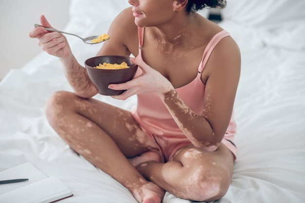 Una donna dalla pelle scura che mangia cereali mentre è seduta sul letto