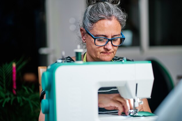 Una donna di mezza età con i capelli bianchi cuce con la macchina da cucire a casa.