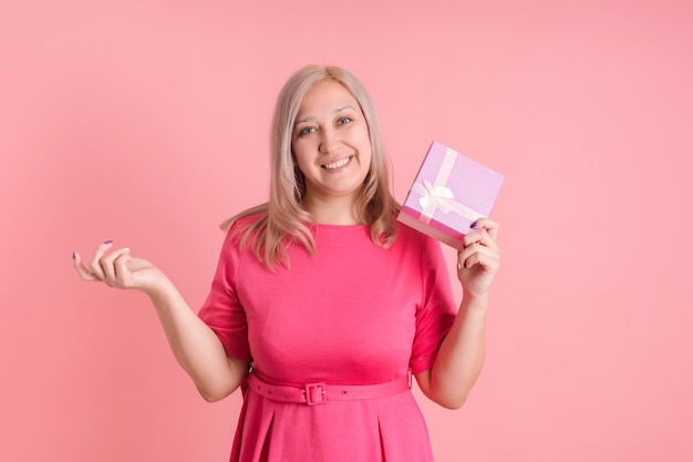 Una donna di mezza età con una confezione regalo in mano si erge su uno sfondo rosa con un vestito rosa