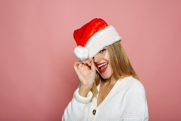 Una donna divertente copre un occhio con un cappello da Babbo Natale e ride. La signora civettuola su uno sfondo rosa celebra il Natale e il Capodanno.