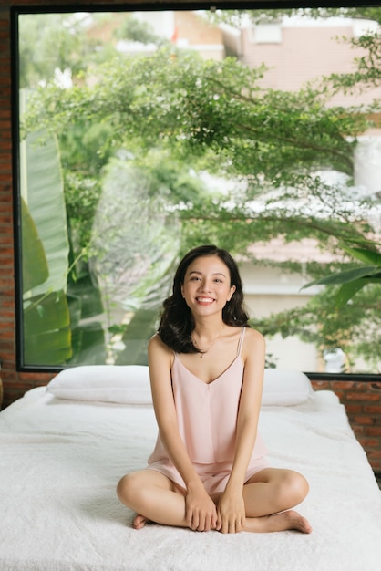 Una donna è seduta sul letto con le gambe piegate, le braccia appoggiate sulle gambe e sorride