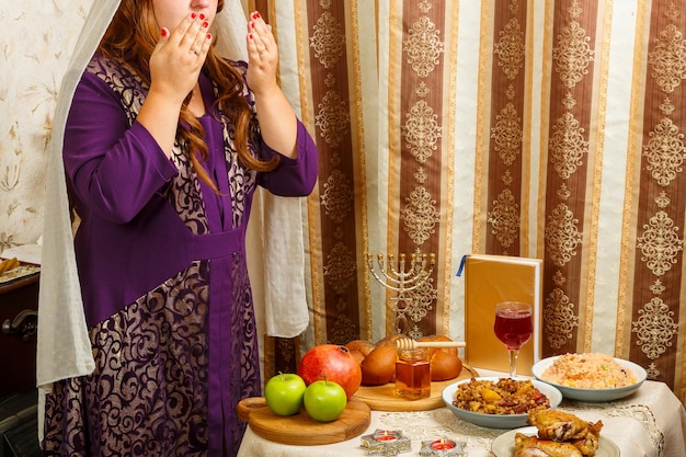 Una donna ebrea con un velo che le cade dai capelli senza volto legge una benedizione sulle candele che le coprono il viso con le mani al tavolo festivo di Rosh Hashanah