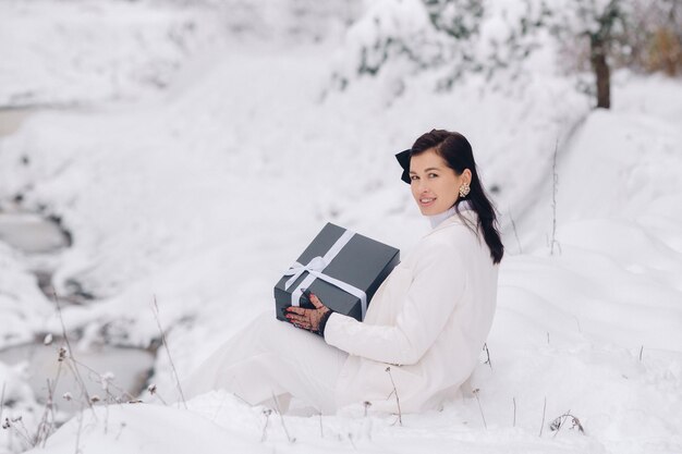 Una donna elegante con un abito bianco con un regalo di Capodanno in mano in una foresta invernale Una ragazza in natura in una foresta innevata con una confezione regalo
