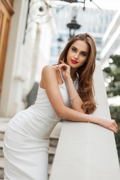 Una donna elegante e elegante con le labbra rosse in un vestito bianco elegante si trova e posa vicino a un edificio vintage